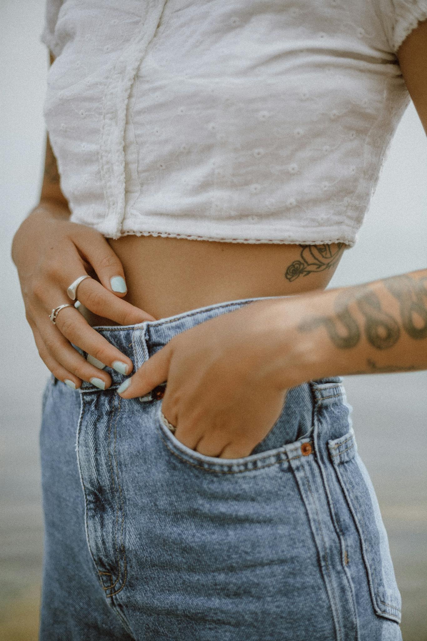 Close-up of a woman's tattooed abdomen wearing a crop top and jeans, showcasing fashion and style.