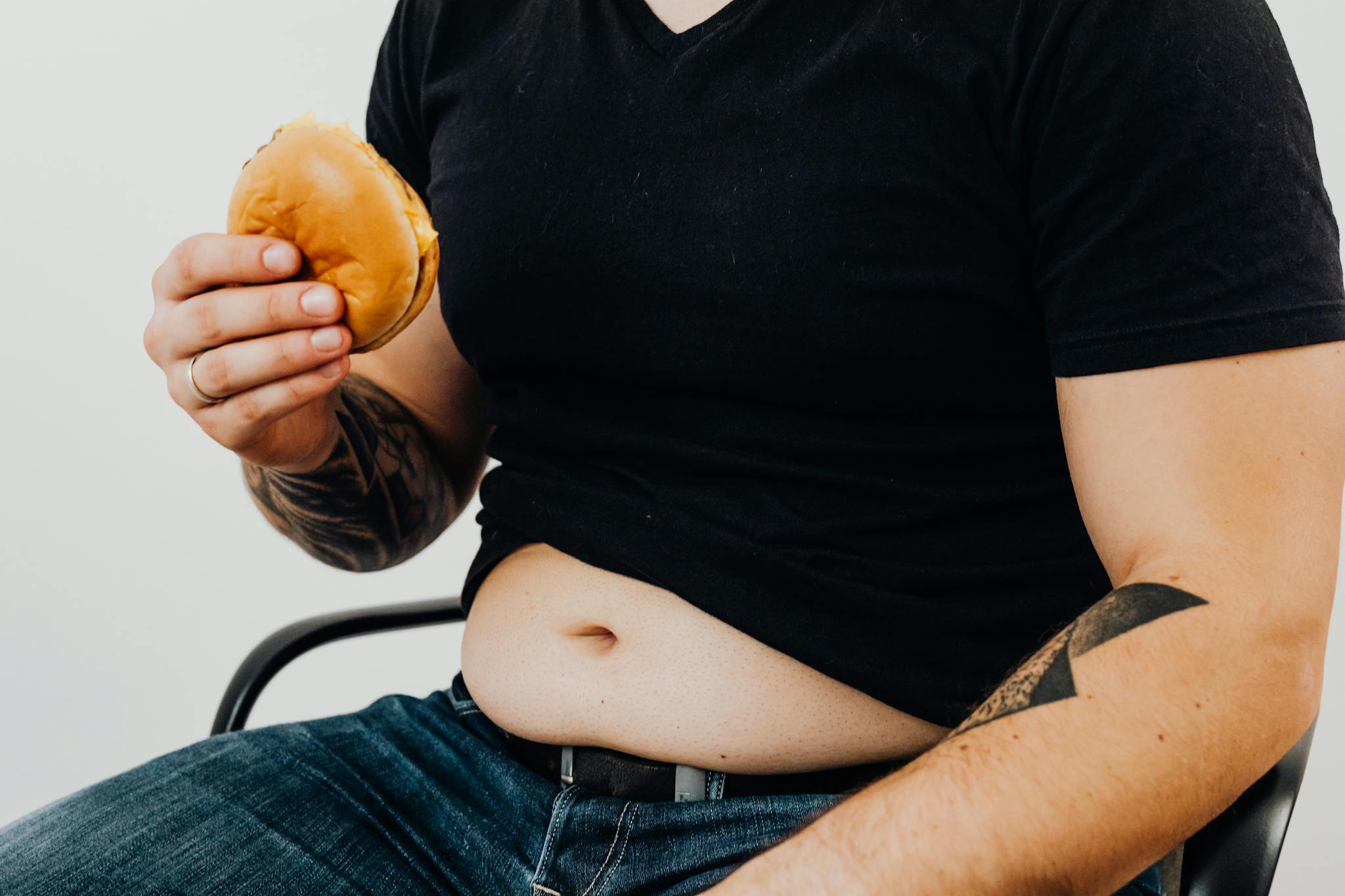 Close-up of a man holding a burger, showcasing his torso and tattooed arm. Casual and relaxed setting.